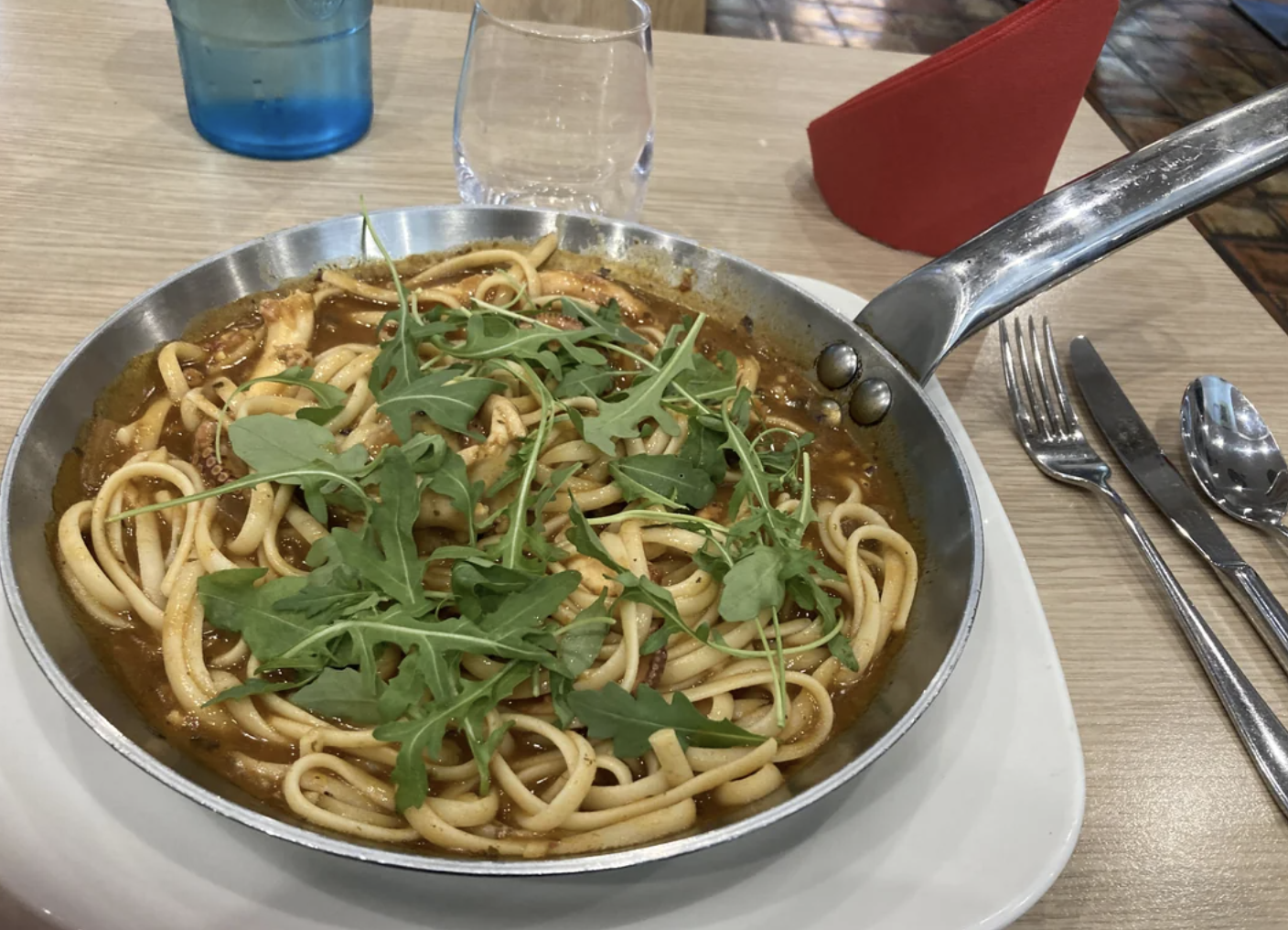 Pasta dish with sauce and herbs served in a pan on a wooden table, with cutlery and glasses on the side