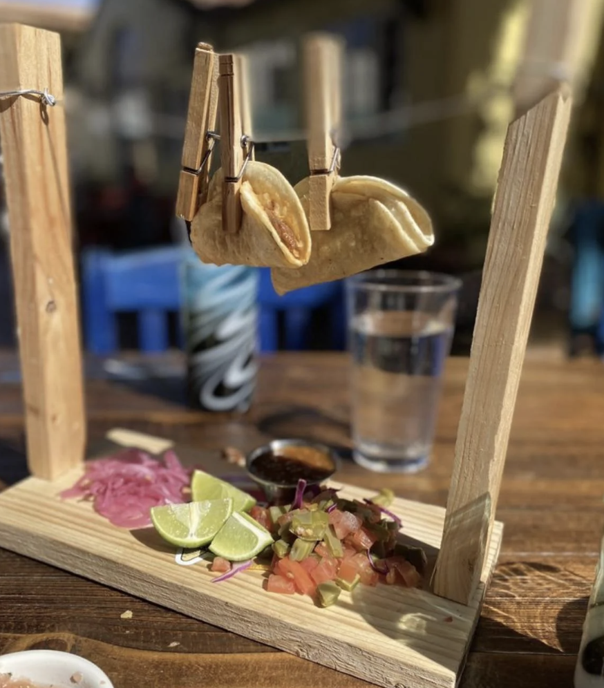 two tacos suspended on a wooden stand with clips, served with lime slices and salsa