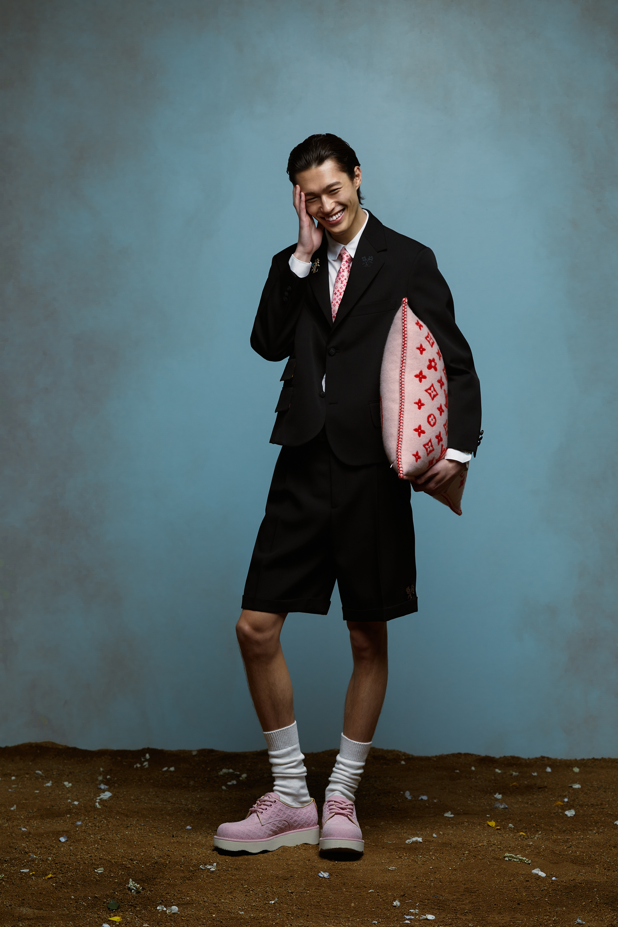 Person in a suit jacket with shorts, holding a patterned pillow, smiling, and touching their temple