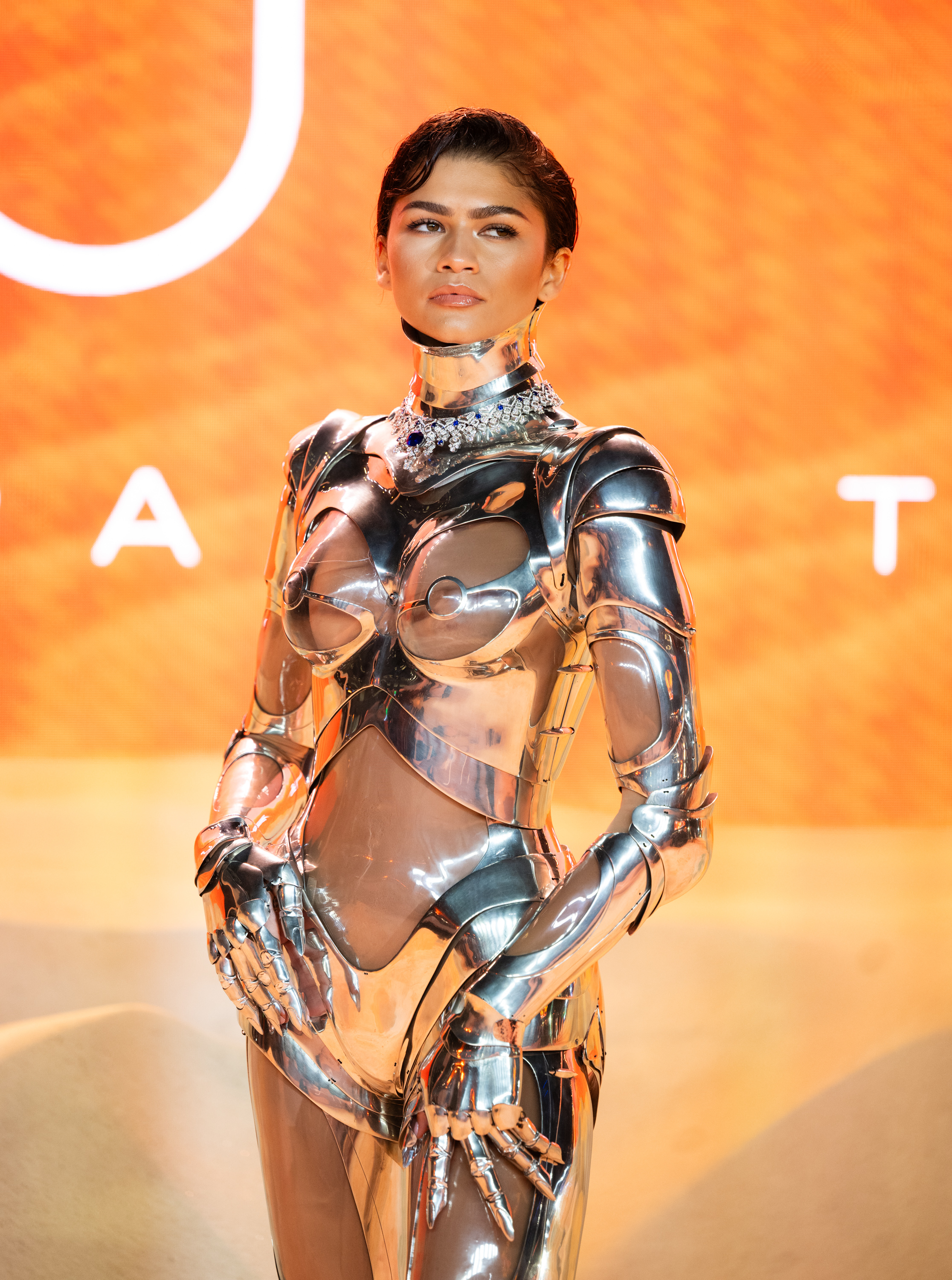 Zendaya in a futuristic metallic outfit posing at an event with an orange backdrop