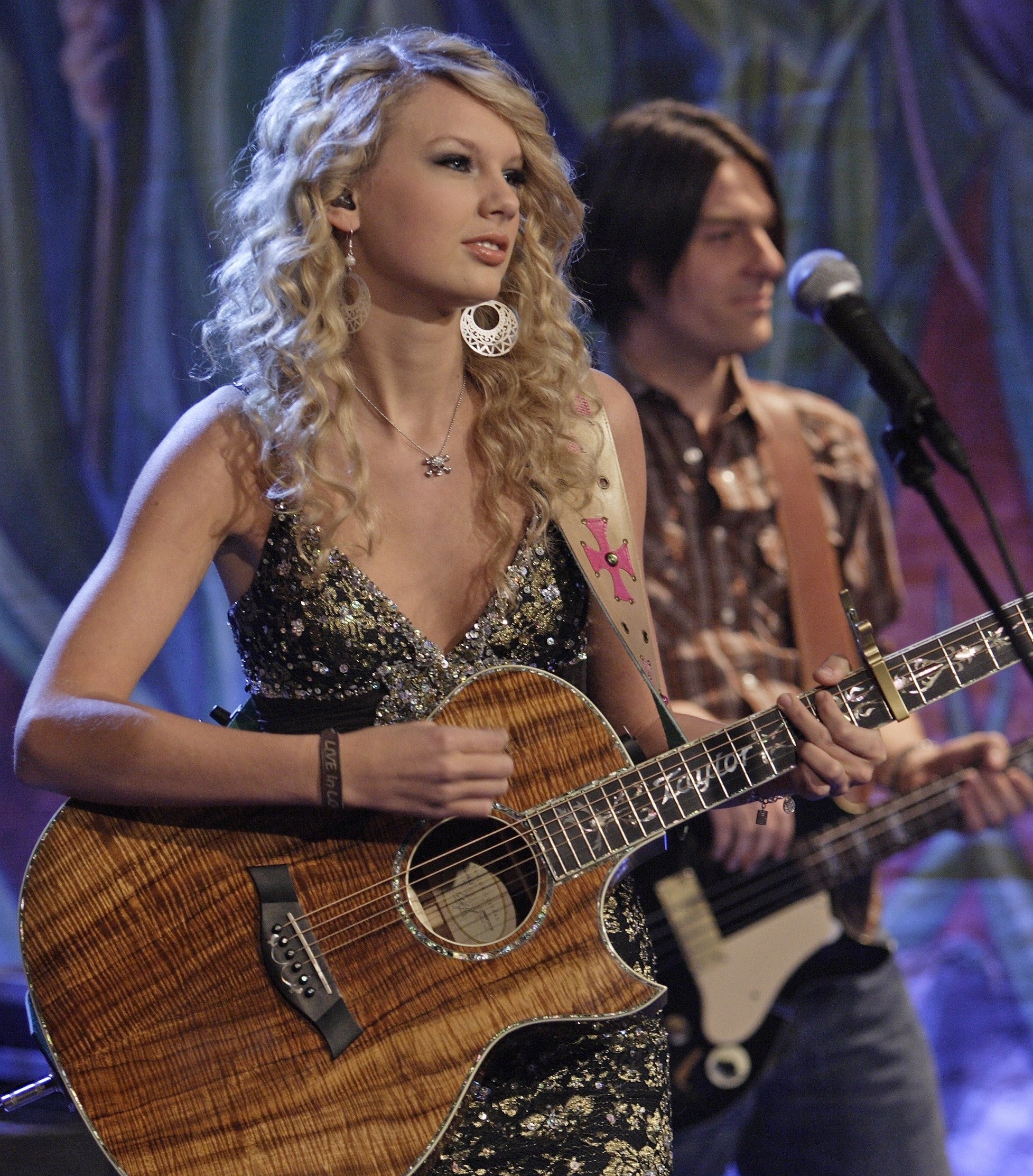 Taylor Swift performing with a guitar, wearing a sparkly black dress, accompanied by a male guitarist