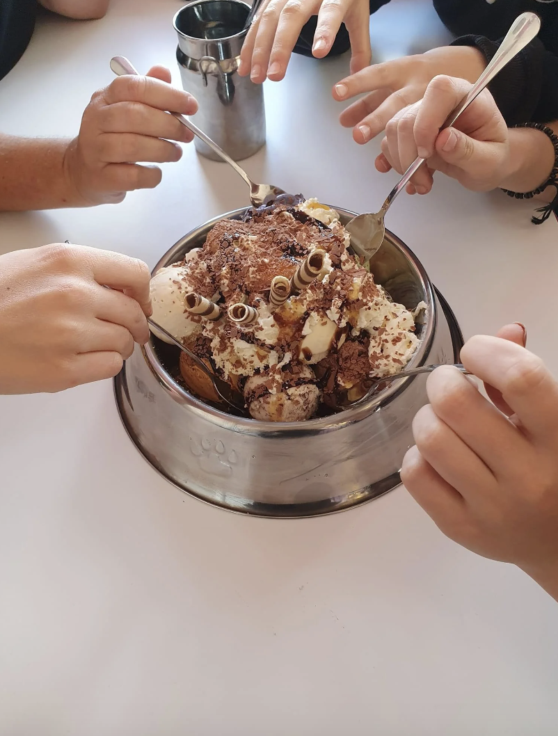 Several hands scooping from a large dog bowl of ice cream with various toppings