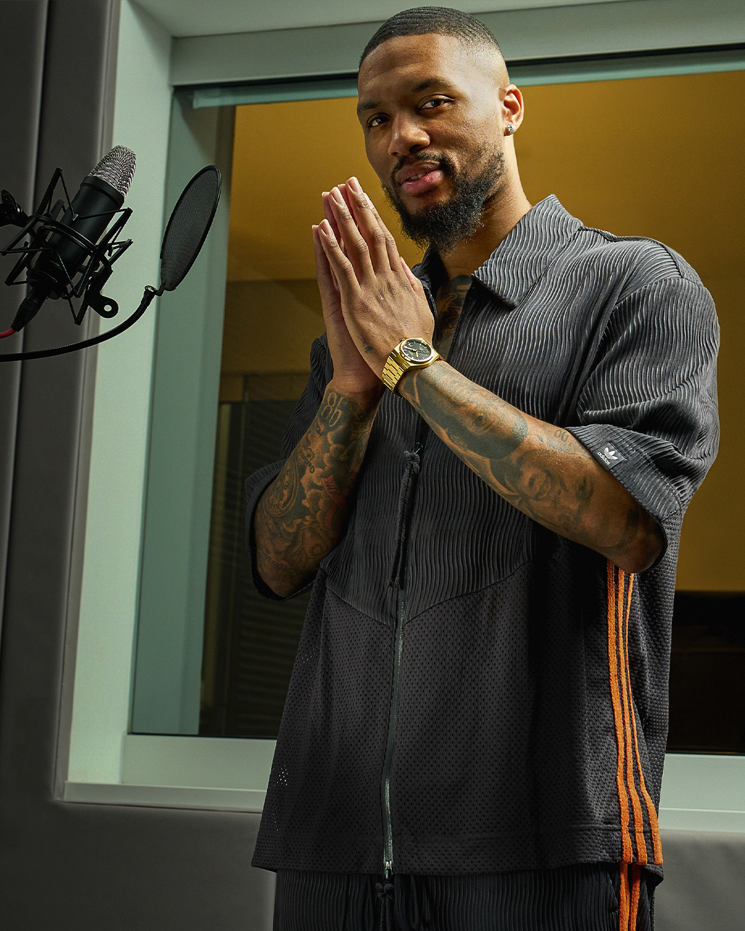 Man in a studio with tattoos visible, standing by a microphone, hands pressed together