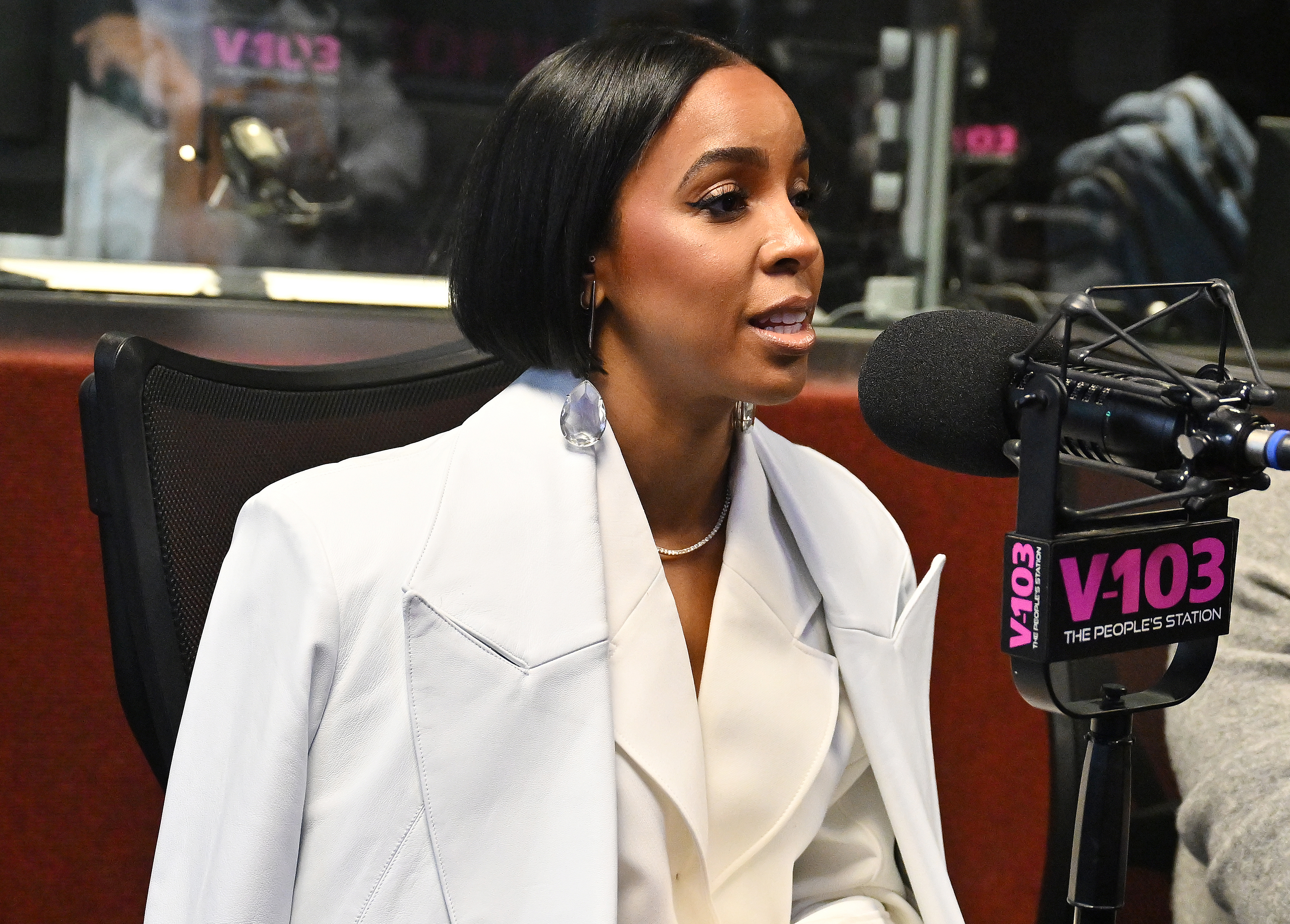 Kelly Rowland in a radio studio, wearing a white blazer, speaking into a microphone