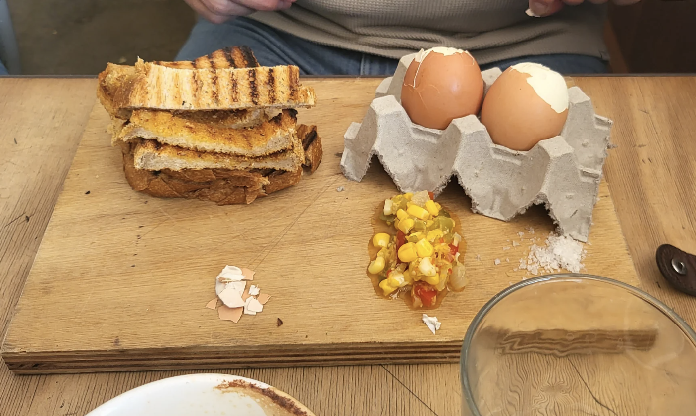 Person at table with a breakfast set, including boiled eggs in their shells and carton, toast, and a side of corn and tomatoes