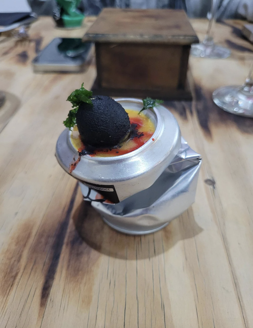 A dish with an edible black sphere garnished with greens on a sauce-covered plate, served on a crushed metal can