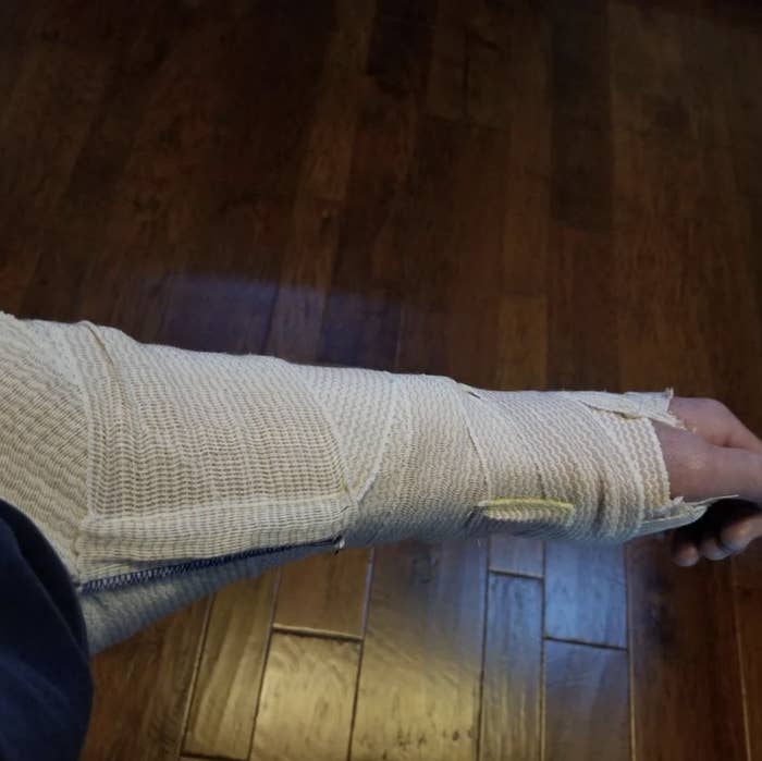 Person&#x27;s arm in a beige medical cast extending outwards with wood flooring in the background