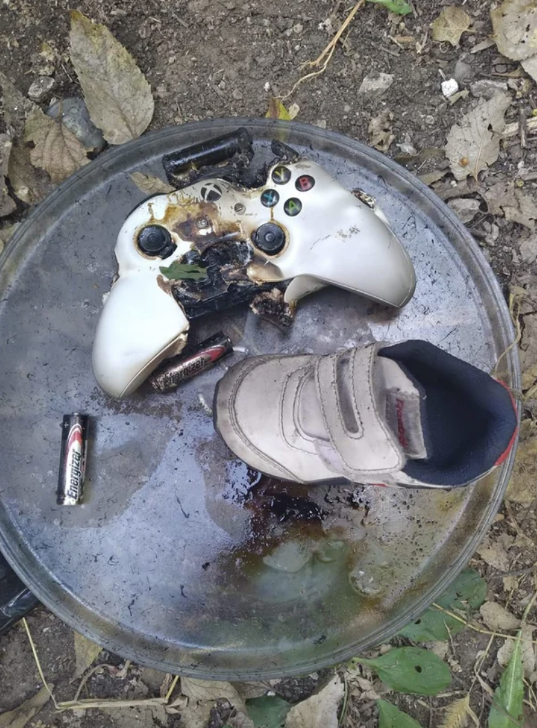 Burnt video game controller and shoe on a metal lid, with batteries and leaf debris
