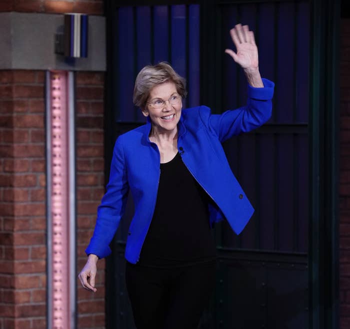 Elizabeth Warren in a blue blazer and black pants, waving on a talk show set