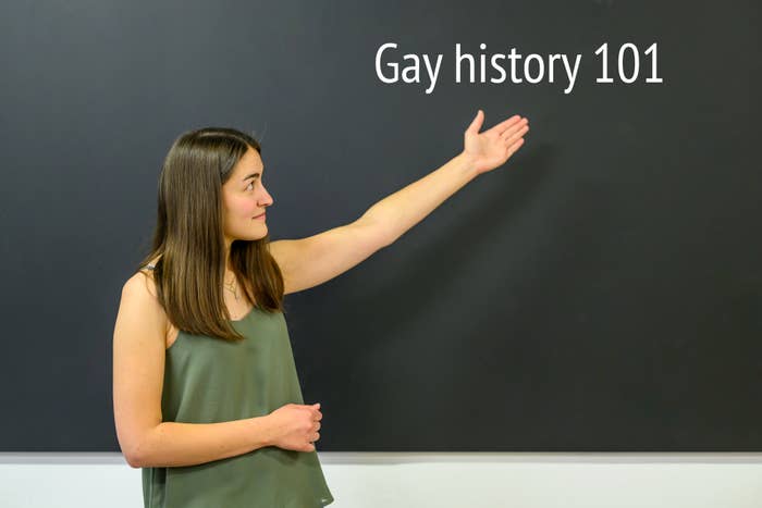 Woman in a sleeveless top gesturing toward a chalkboard with &quot;Gay history 101&quot; on it
