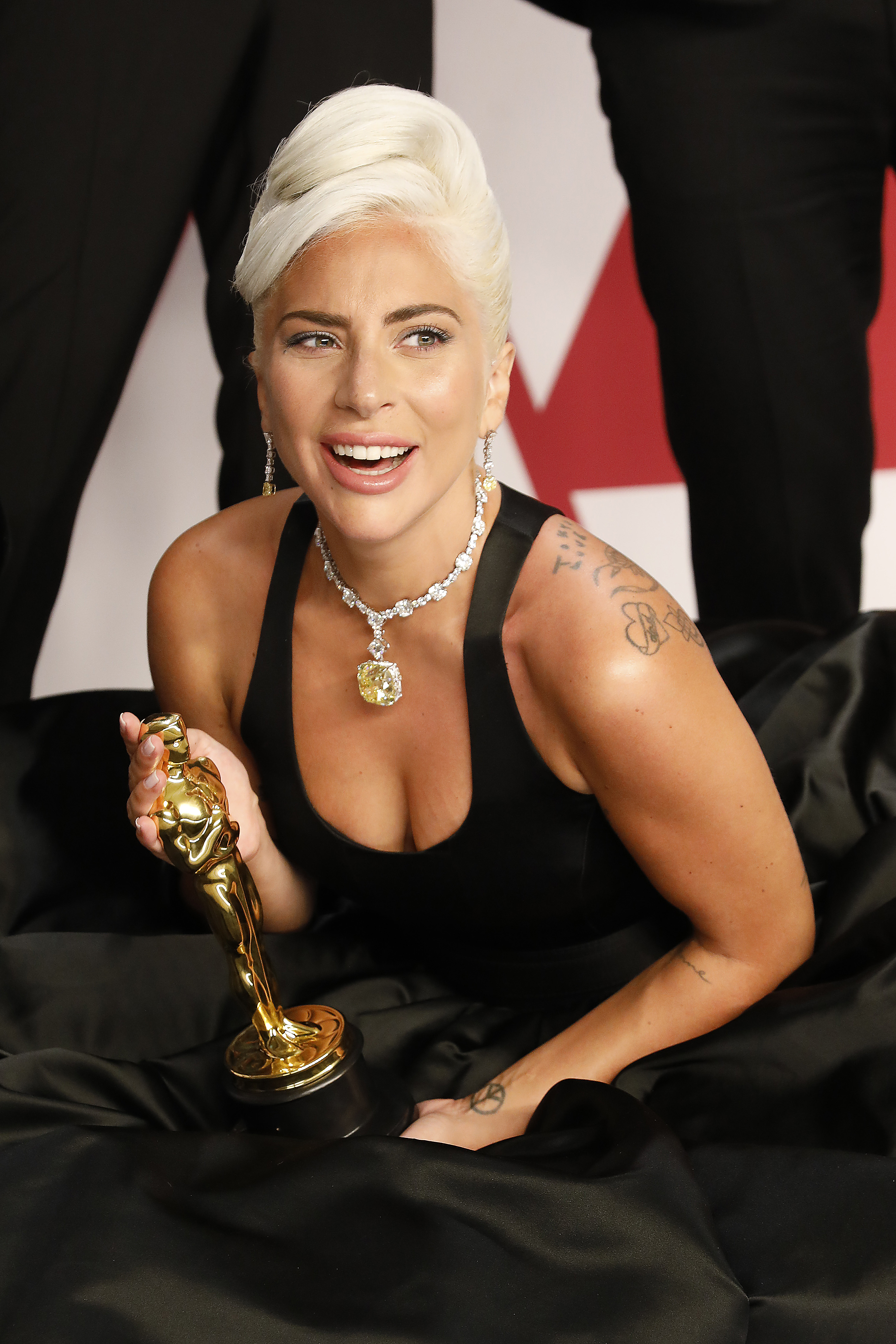Lady Gaga in a black dress holding an Oscar trophy, smiling, with a necklace