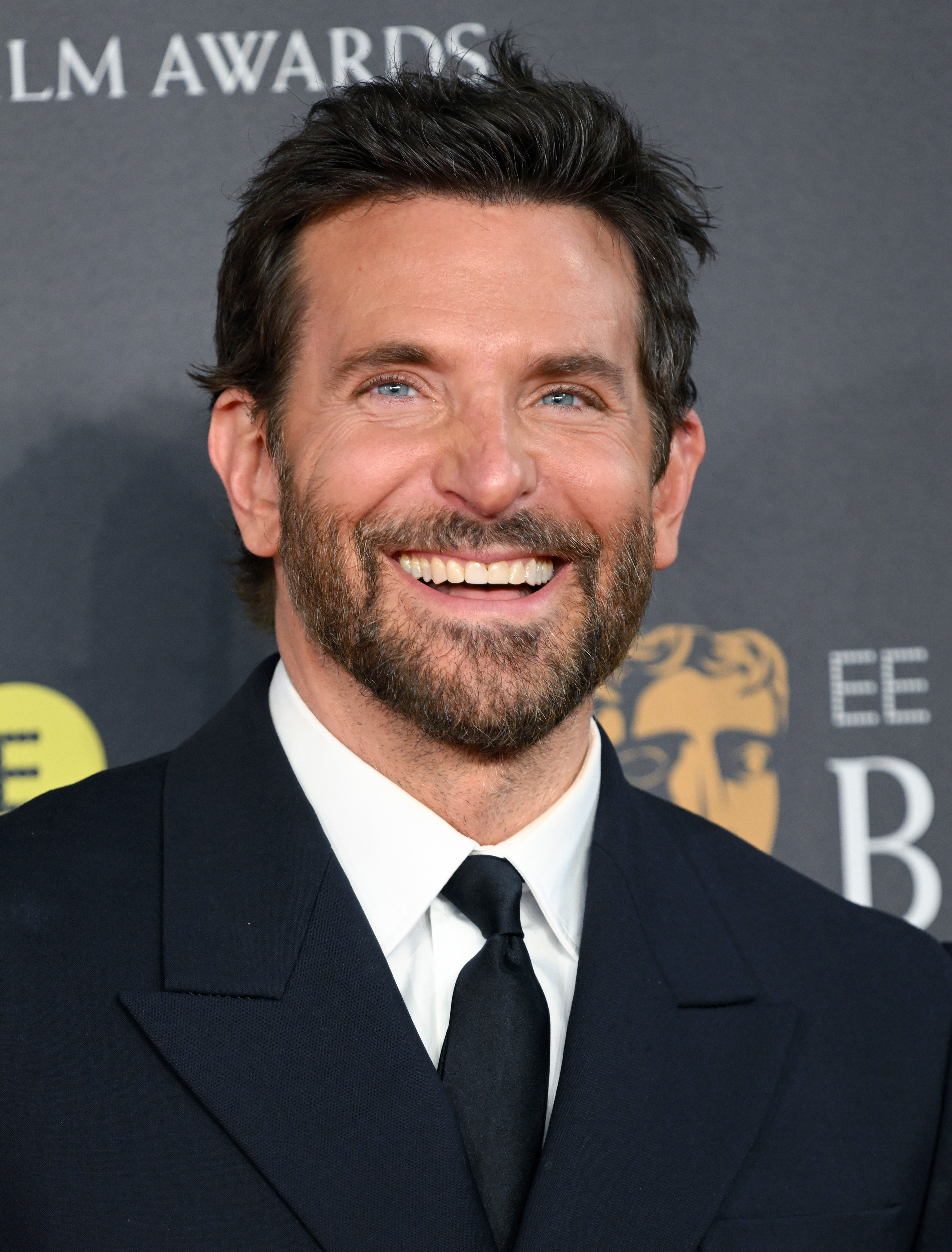 Bradley smiling in a classic black suit with a tie at a media event