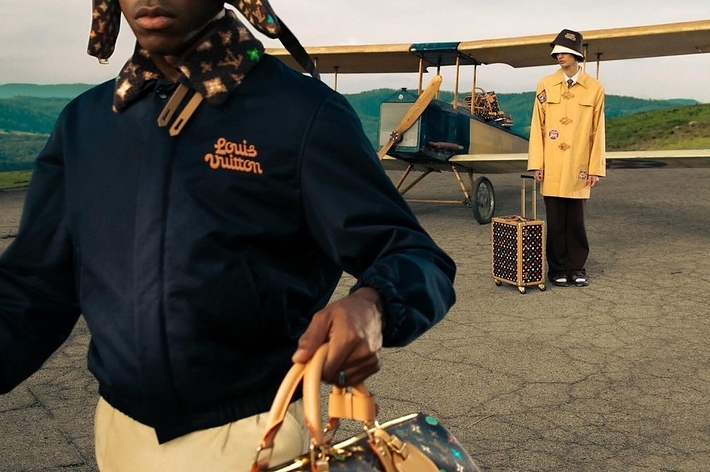 Two models in vintage pilot attire with Louis Vuitton bags, one foreground holding a duffel, the other by a plane