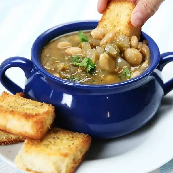A hand dipping a toasted bread slice into a blue pot of bean soup with vegetables