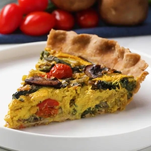 A slice of vegetable quiche on a white plate with tomatoes and mushrooms in the background