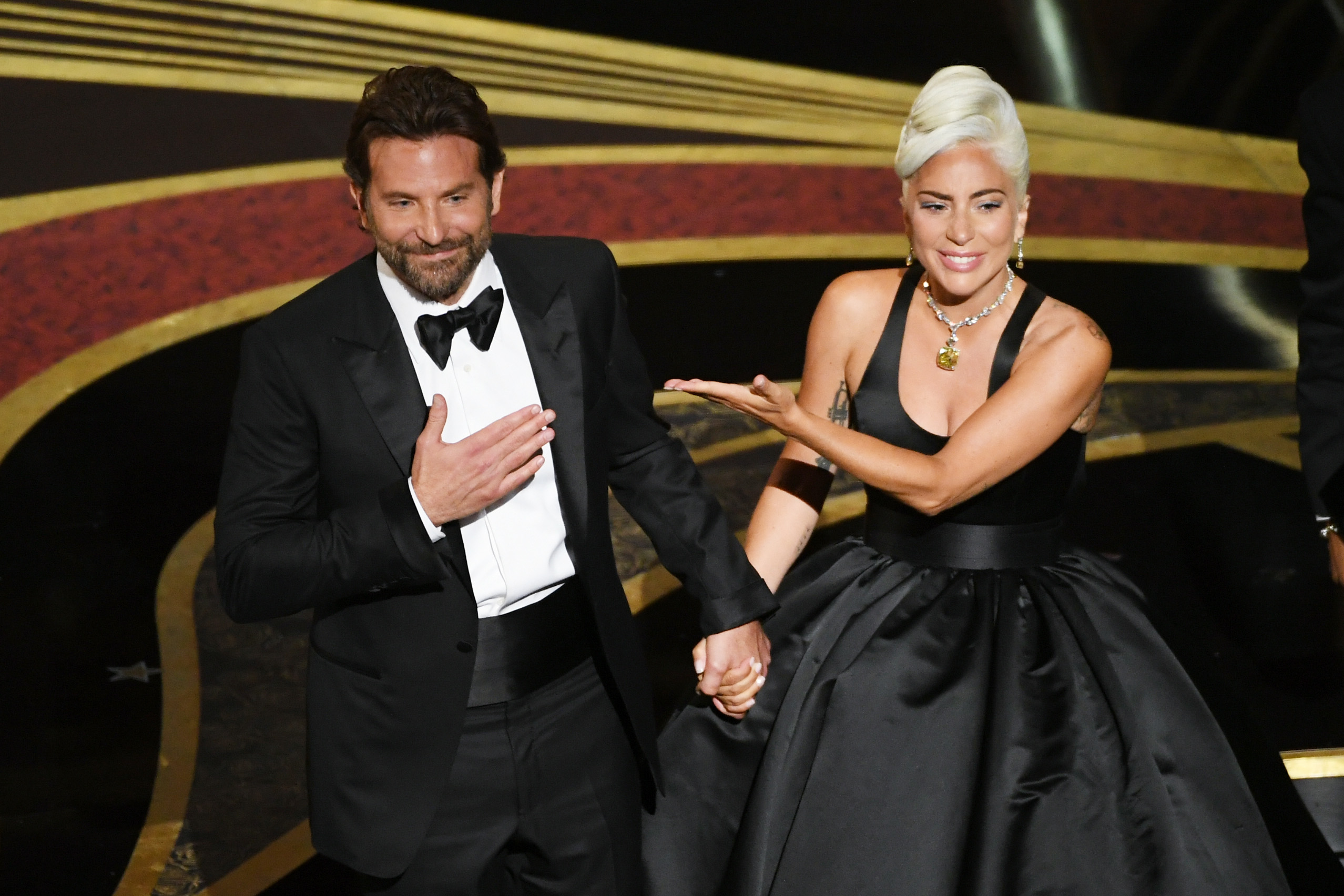 Bradley Cooper and Lady Gaga onstage holding hands; he&#x27;s in a tuxedo, she&#x27;s in a black gown with a prominent necklace