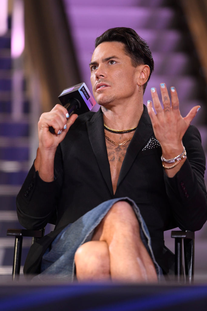 Tom in an open jacket speaks into a microphone, wearing multiple rings and a necklace