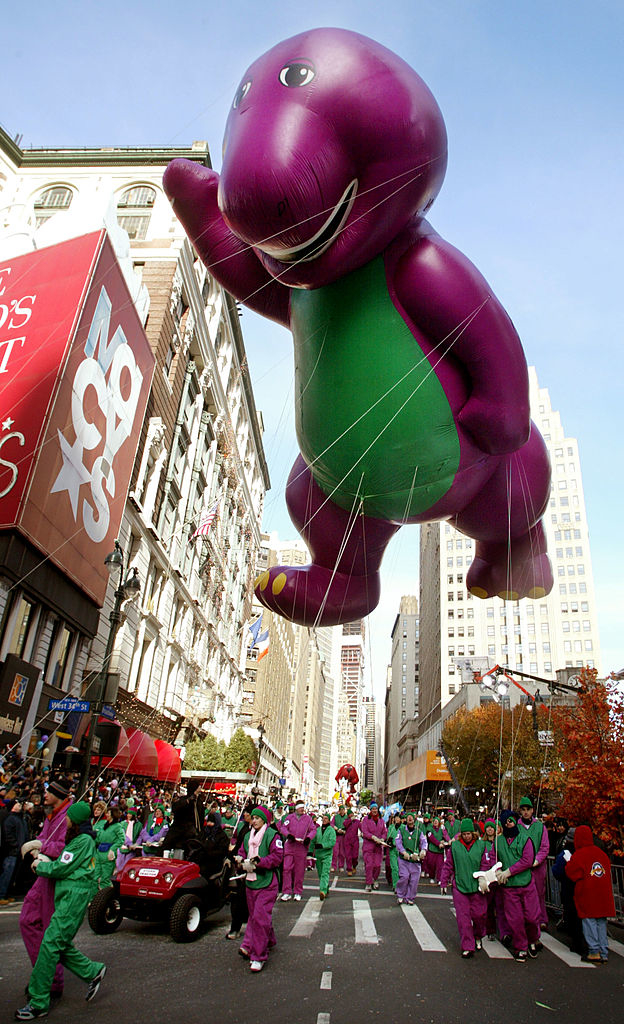 Giant Barney balloon floats in parade with handlers in green uniforms guiding it down a NYC street