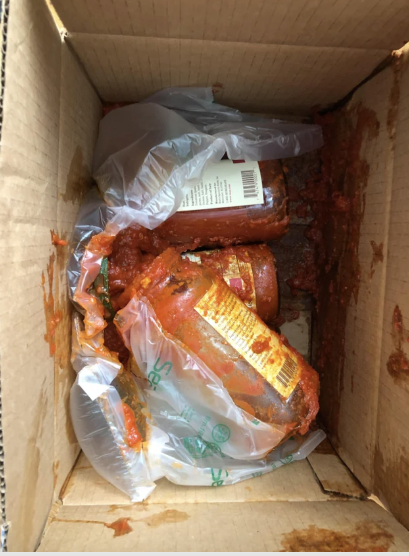 A damaged package with spilled tomato sauce and food items inside a cardboard box