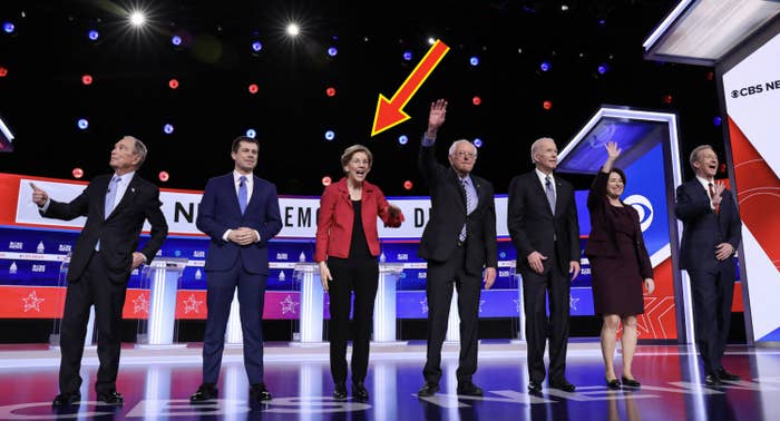 Seven political figures stand on a debate stage with podiums, waving to the audience during a televised event