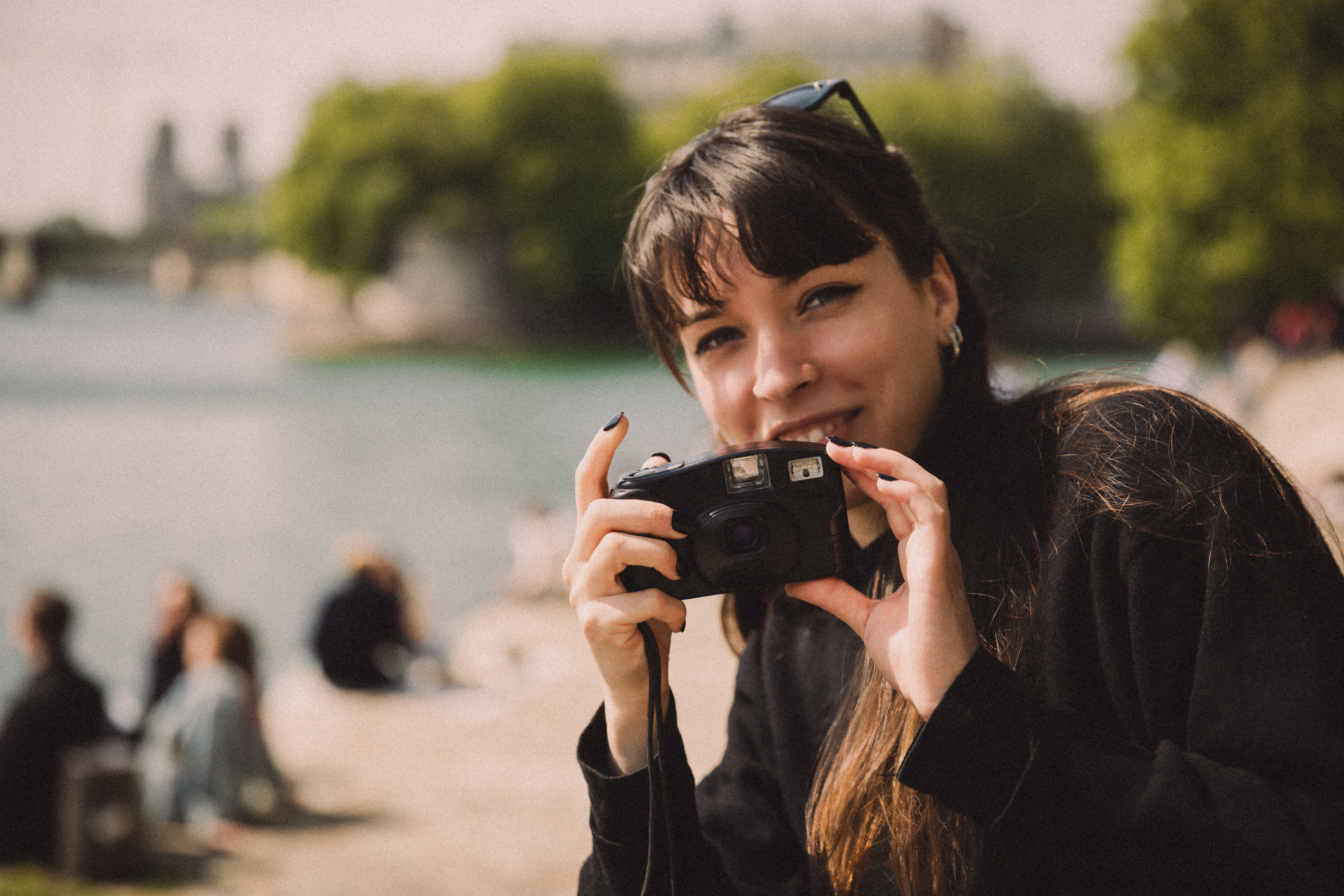 Woman smiling at the camera holding a camera, outdoors with people in the background
