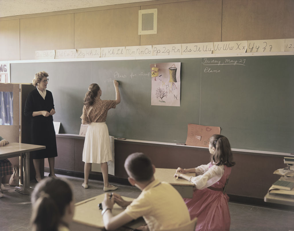 1950s classroom scene with students seated and a girl writing on the chalkboard as the teacher observes