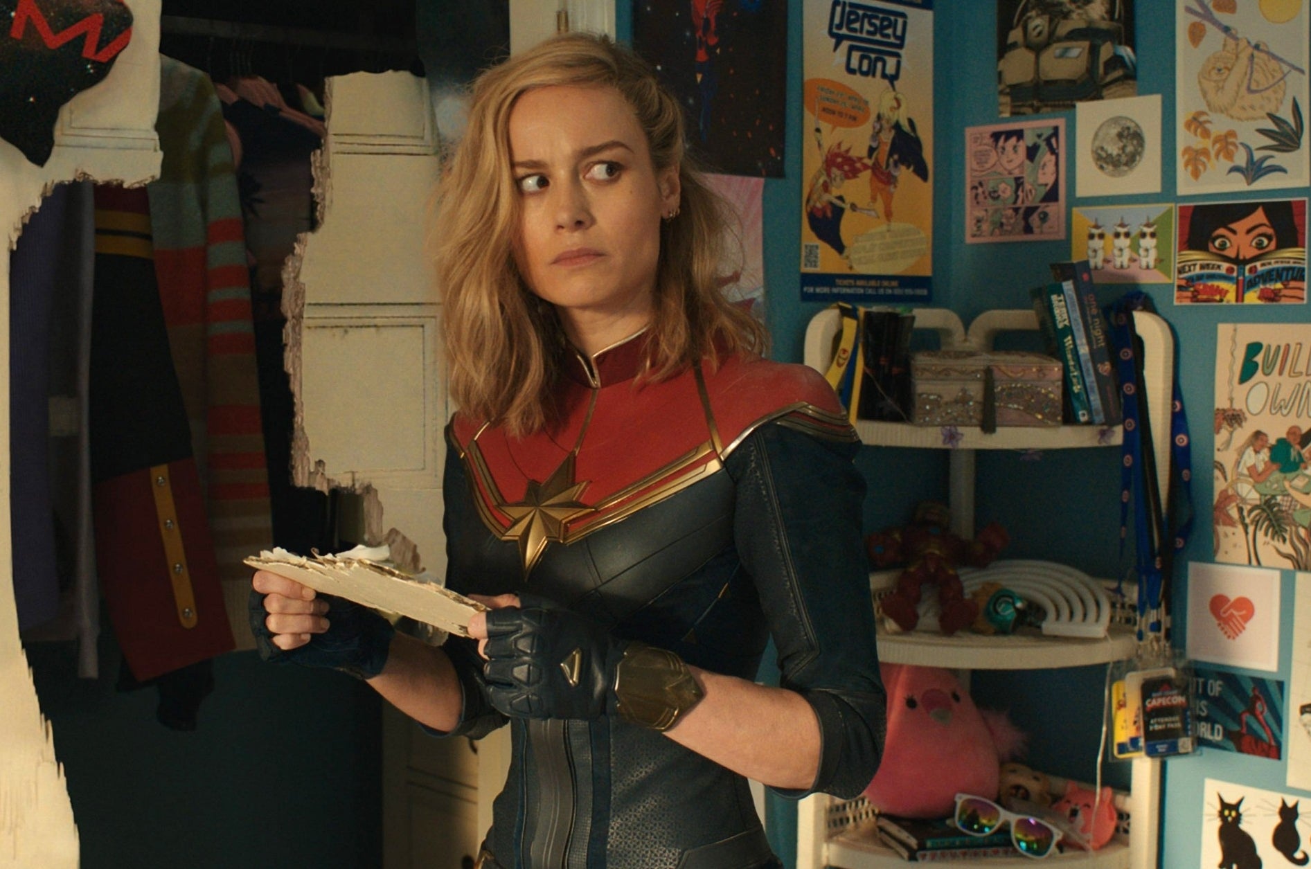 Carol Danvers in a room with vibrant posters, looking pensive while holding a slice of pie