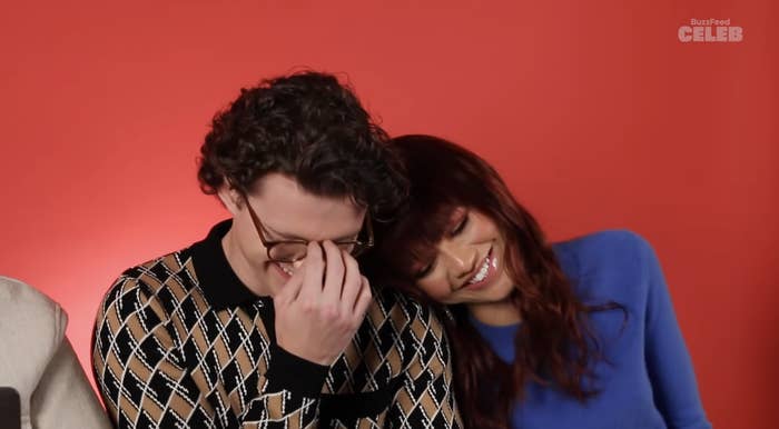 Tom, wearing a checkered top, and Zendaya, who is resting her head on his shoulder, laughing