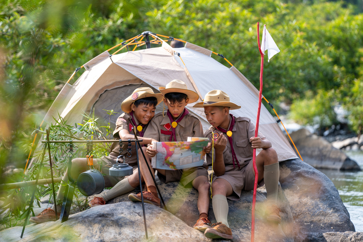 Three children dressed as scouts with a tent by a river, looking at a map and smiling