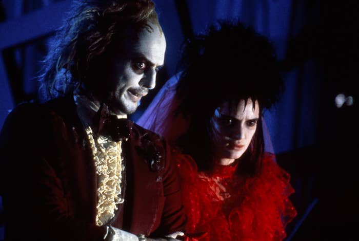 Beetlejuice in a ruffled shirt and black suit, Lydia in a red tulle dress, both with pale faces and dark eye makeup
