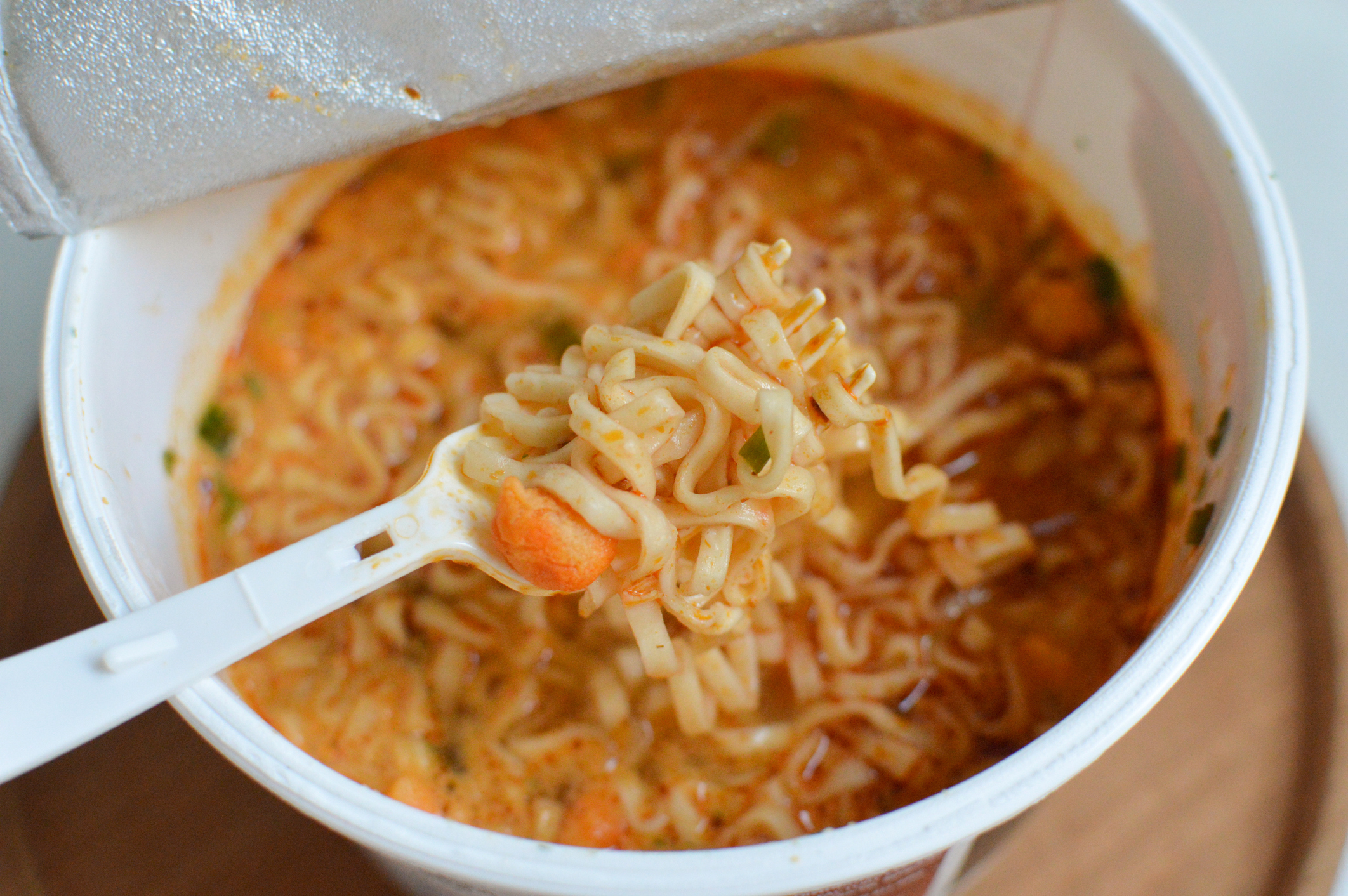 A close-up of a hand holding a fork with instant noodles; a cup of noodles is visible below