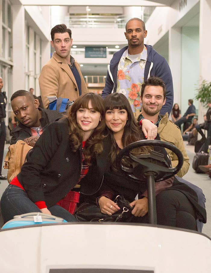 A group of friends from the TV show "New Girl" sitting together, one in a luggage cart, in a busy airport setting