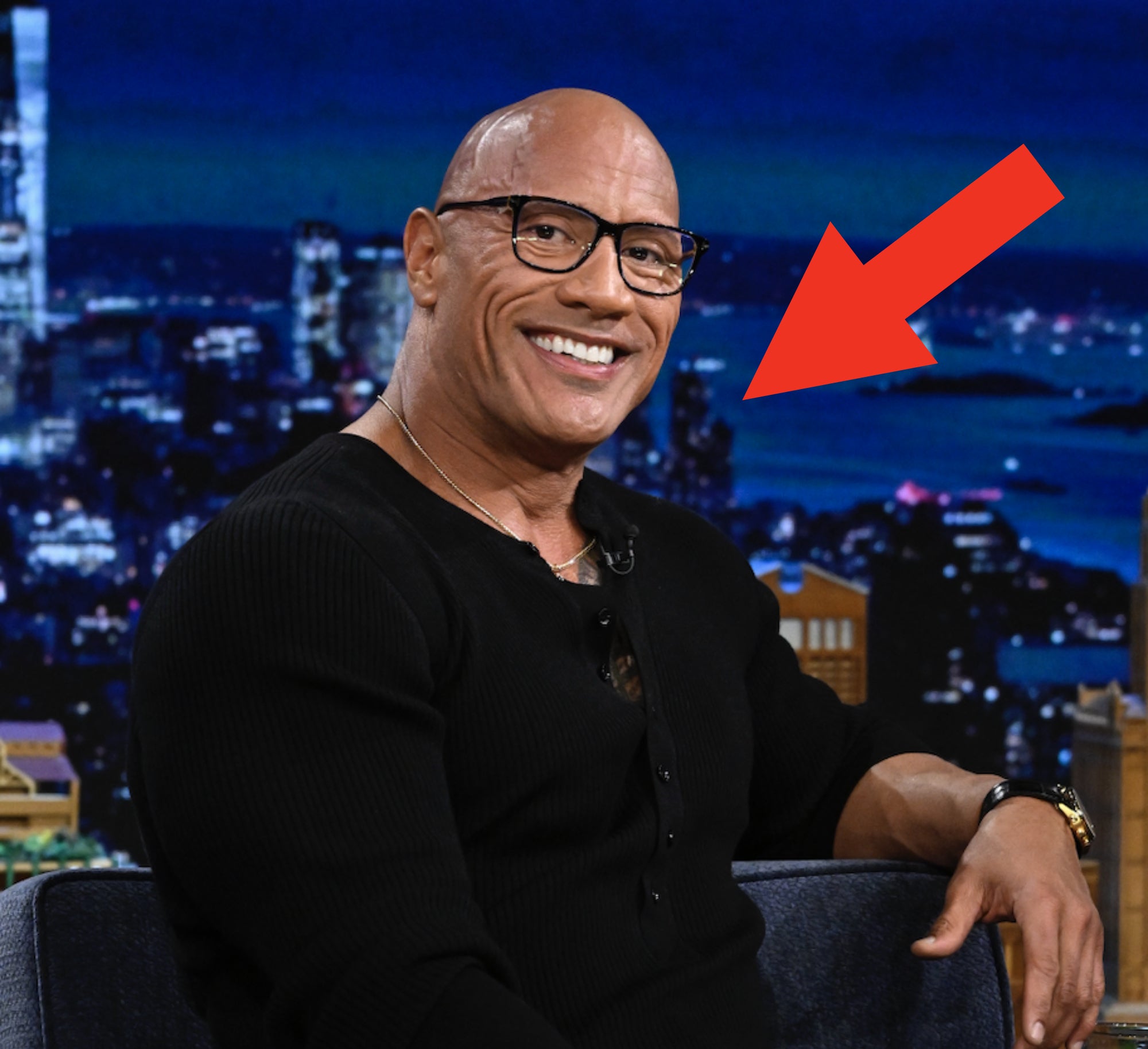Dwayne Johnson seated on a talk show set, smiling, in a black outfit with wrist accessories