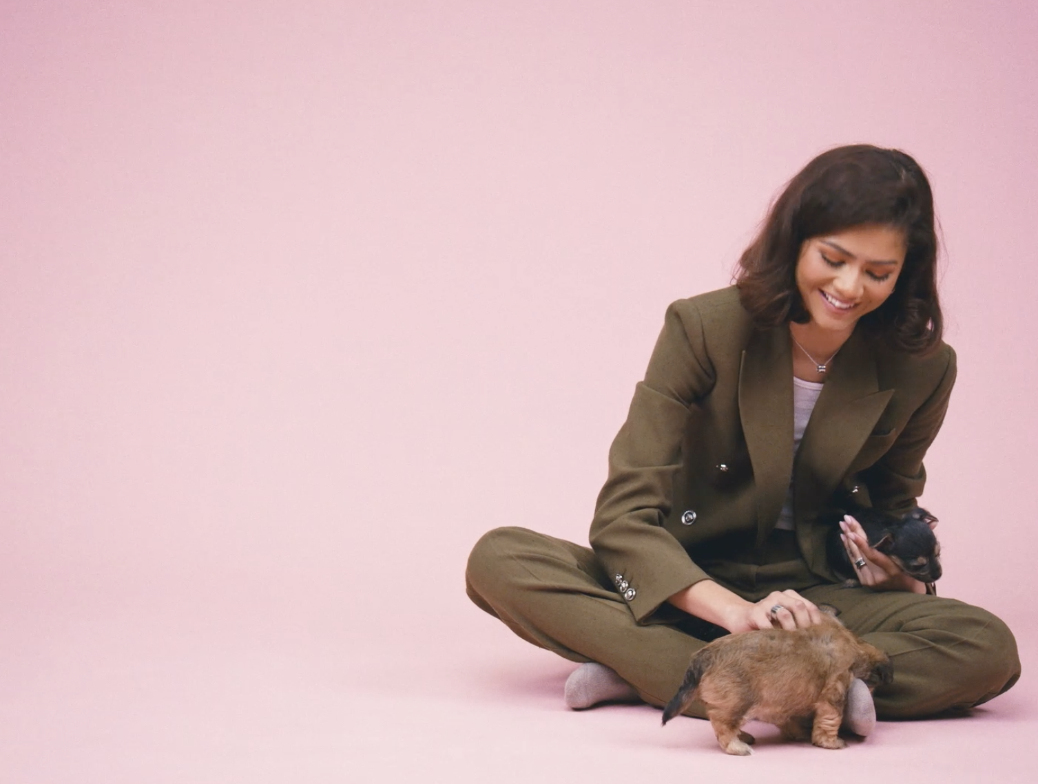 Zendaya, in a pantsuit, kneels and pets a puppy on a pink backdrop, with &quot;Buzzfeed CELEB&quot; logo in the corner