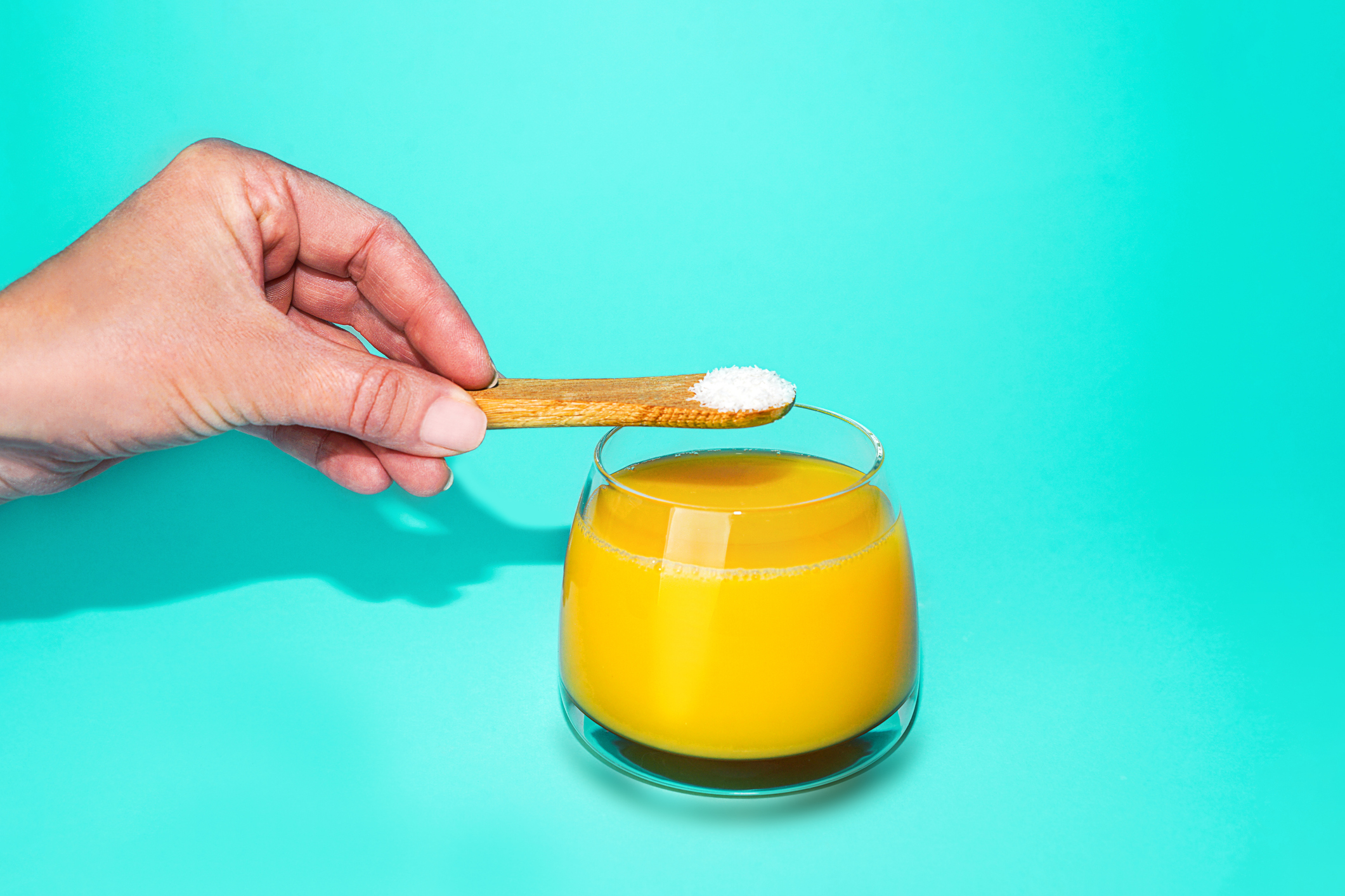 Hand adding a pinch of salt to a glass of orange juice on a turquoise background