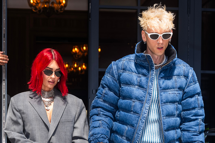 Megan Fox and Machine Gun Kelly exit a building. Fox in a gray suit, MGK in a denim jacket