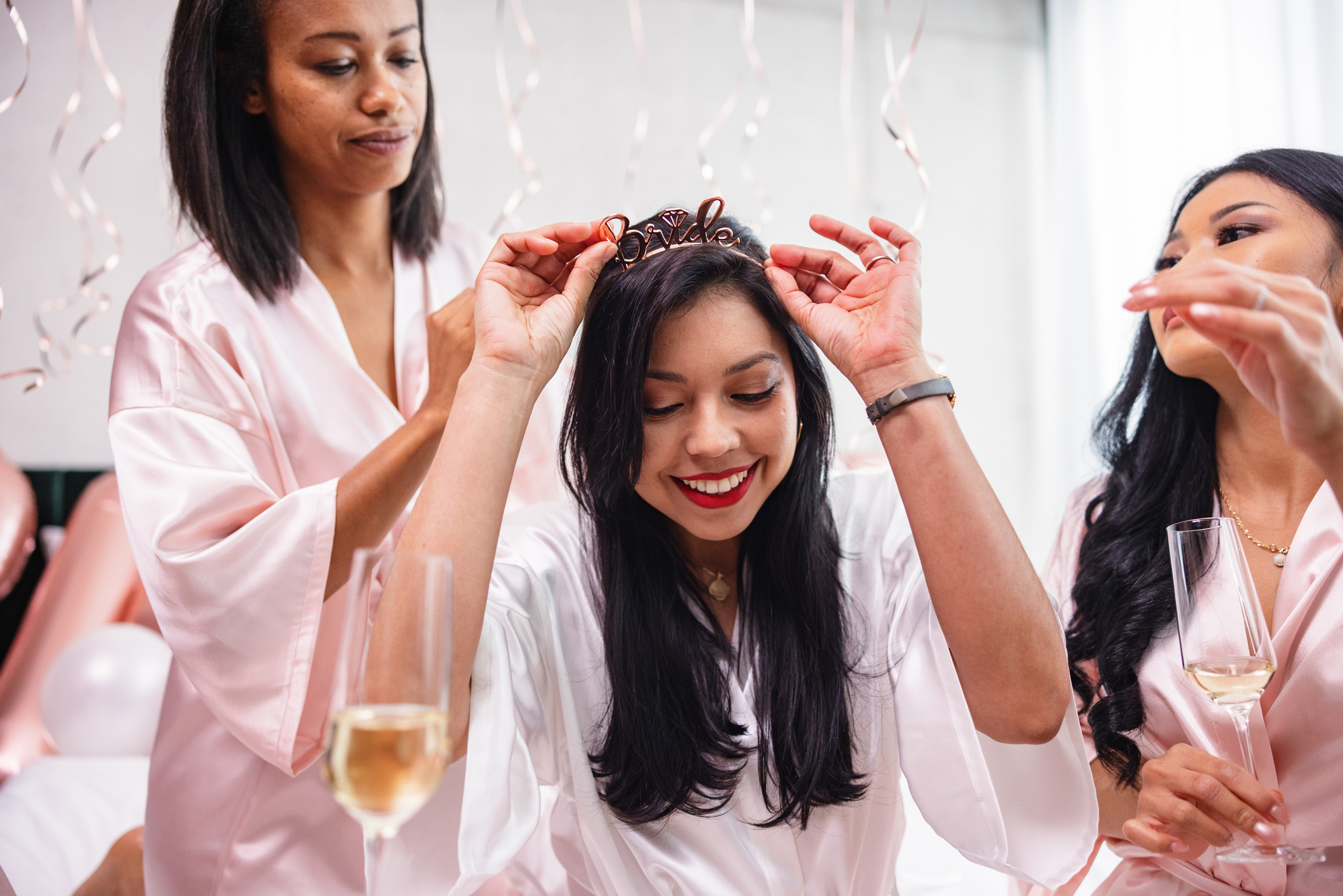 Three women celebrate, one adjusting a tiara, with drinks nearby at a baby shower