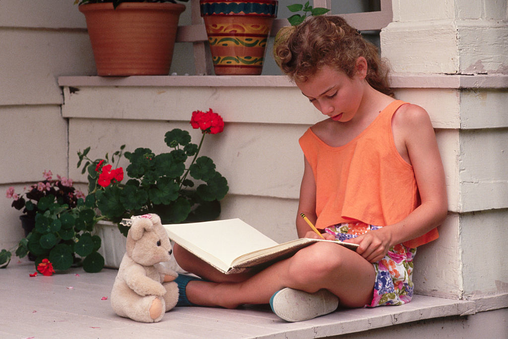 Young girl sits on steps, focused on writing in a book, with a plush toy beside her