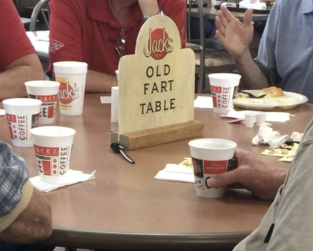 A group of seniors socializing at a table with a humorous &quot;Jack&#x27;s Old Fart Table&quot; sign