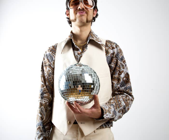 Person in a patterned shirt and vest holding a disco ball, wearing sunglasses and posing confidently