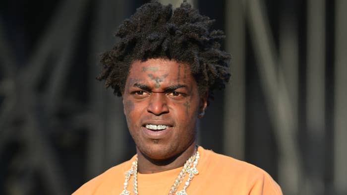 Rapper with dreadlocks and facial tattoos wearing chains and an orange shirt, posing for the camera