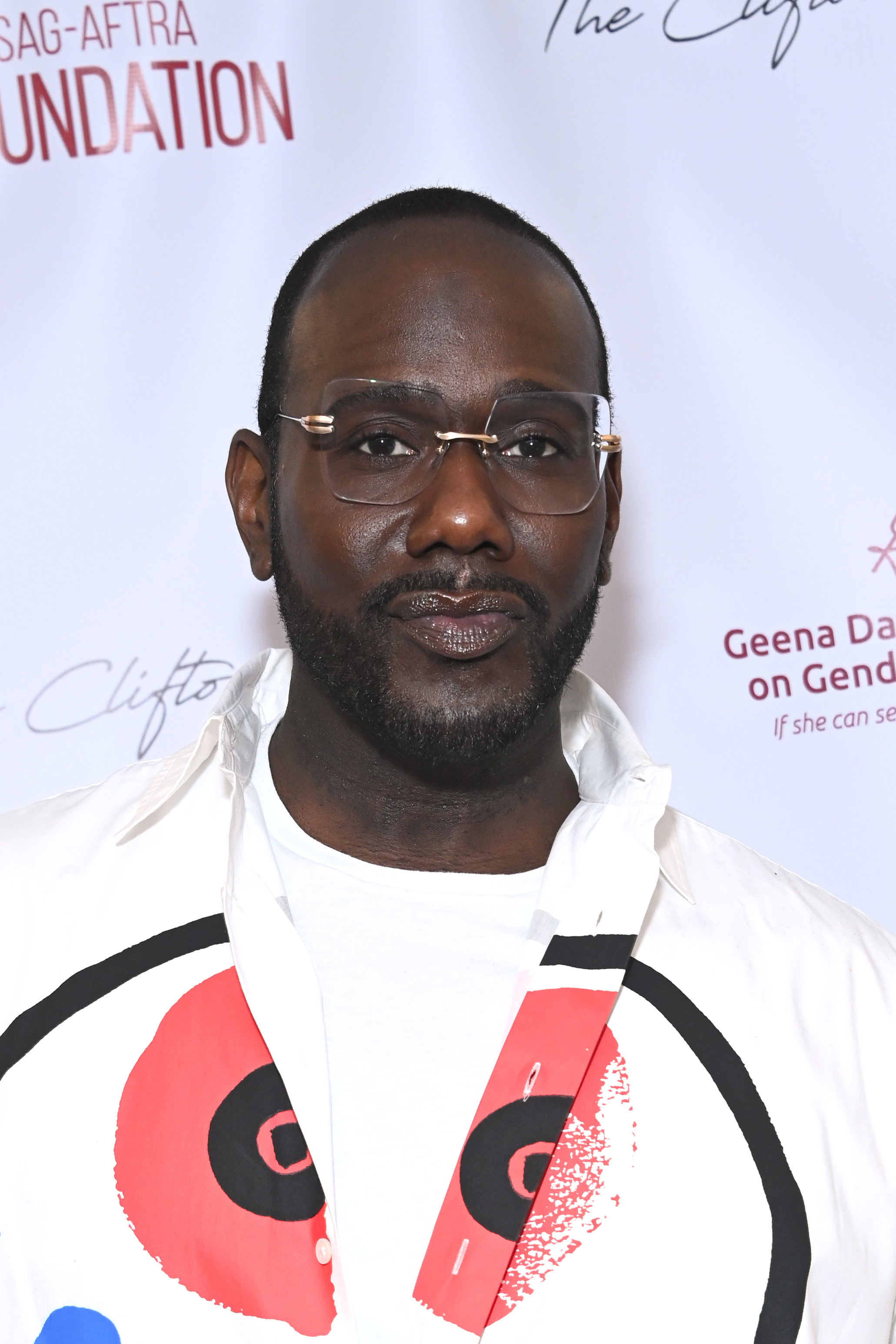 Man in unique glasses and white shirt with graphic design, standing before a promotional backdrop