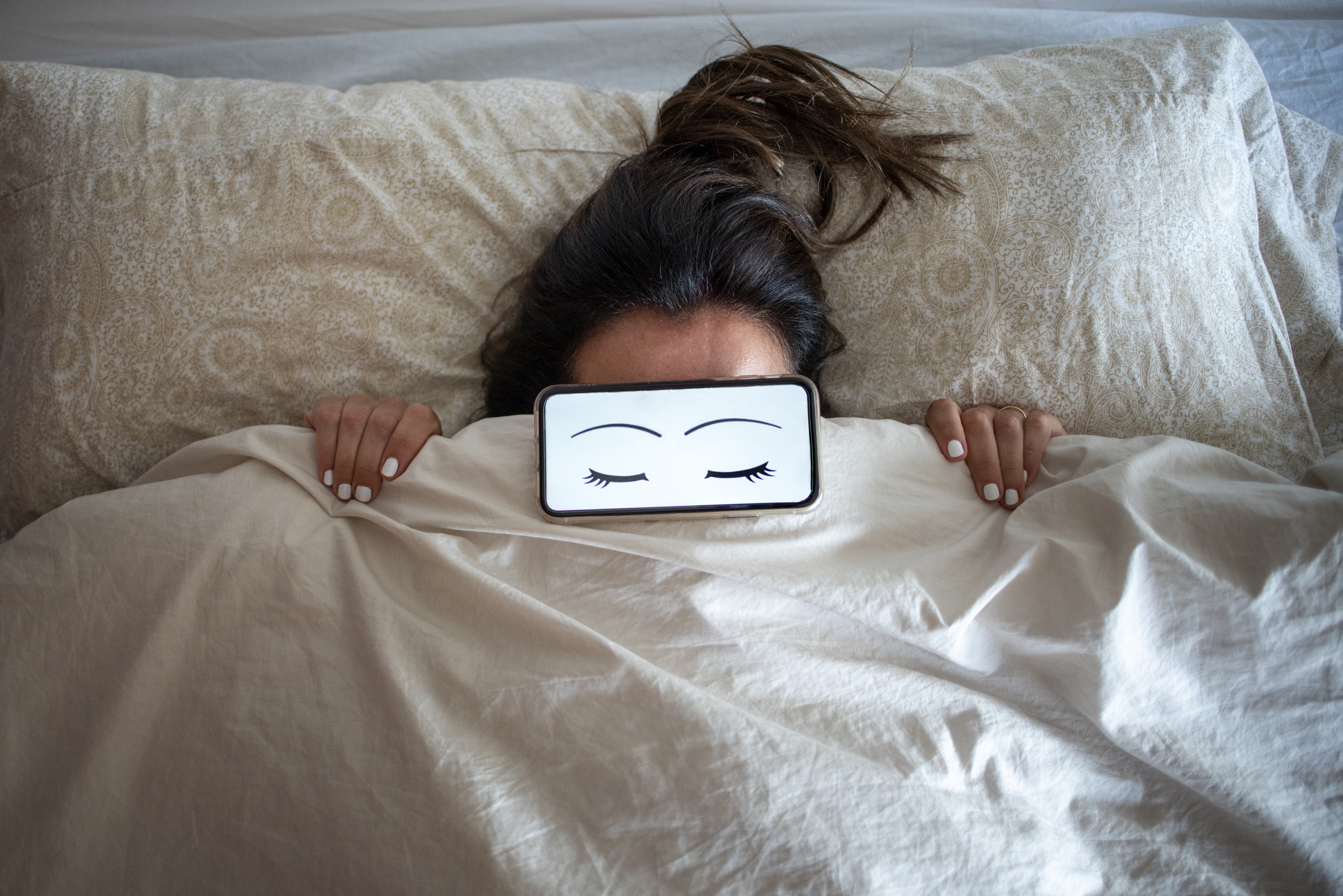 Person lying in bed covered up to forehead with a beige blanket, holding a smartphone over face displaying closed-eye graphic