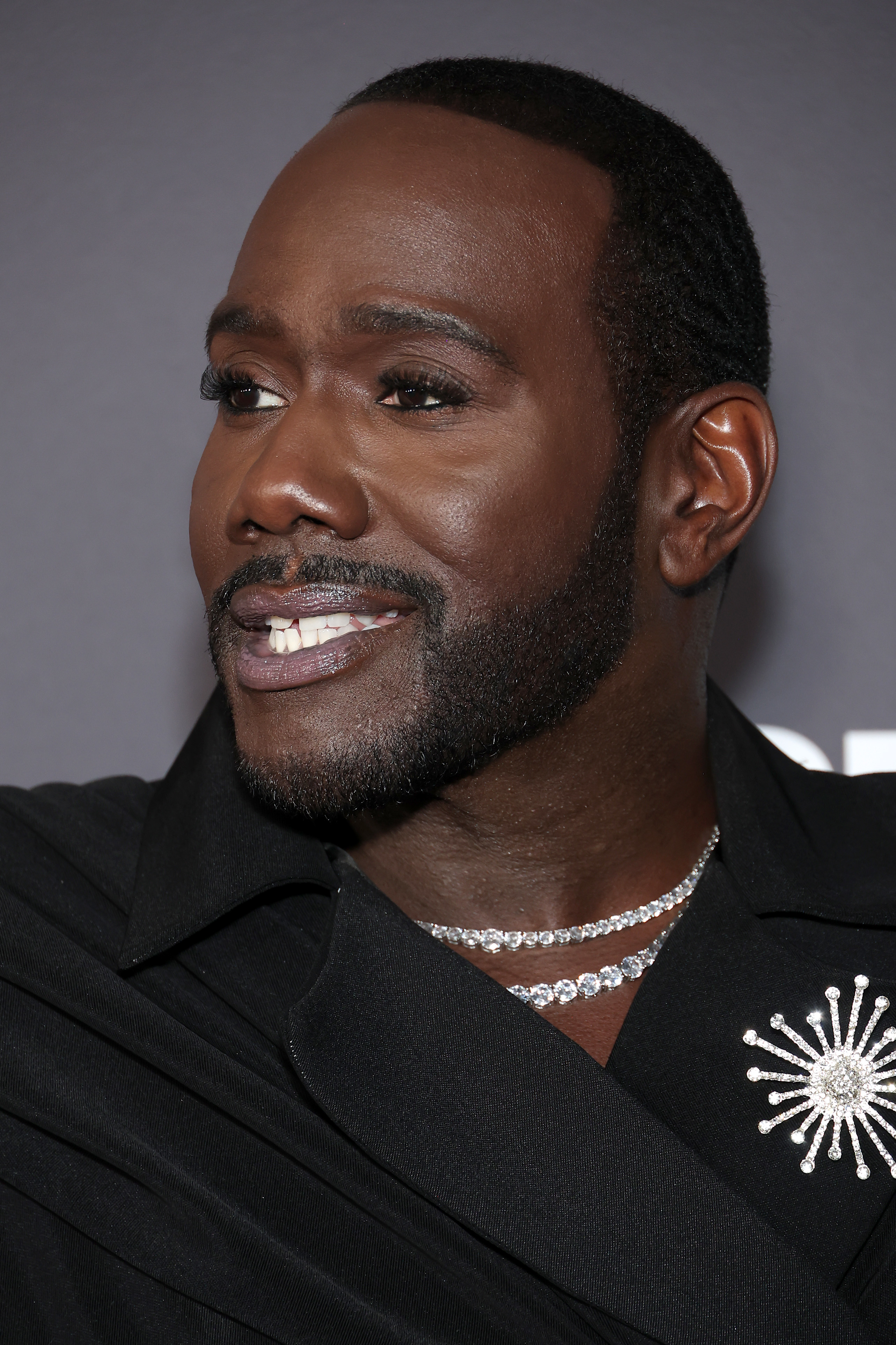 Close-up of a celebrity man smiling, wearing a black jacket with a decorative pin and a diamond necklace