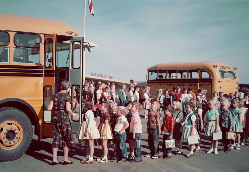 Children lining up to board school buses, supervised by an adult; vintage setting