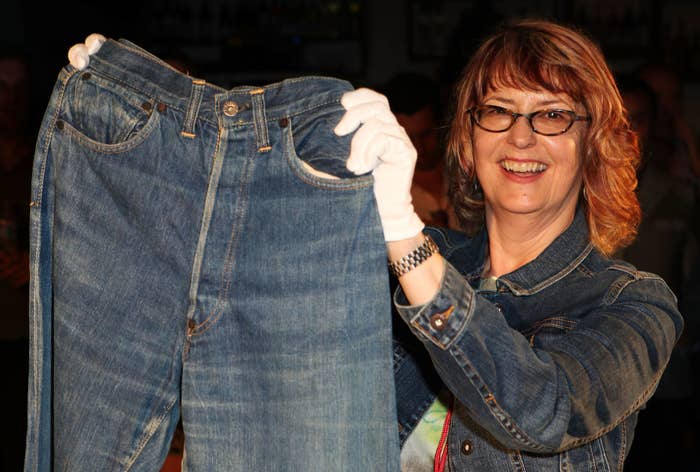 Woman holding up a large pair of jeans, smiling. She wears glasses and a denim jacket