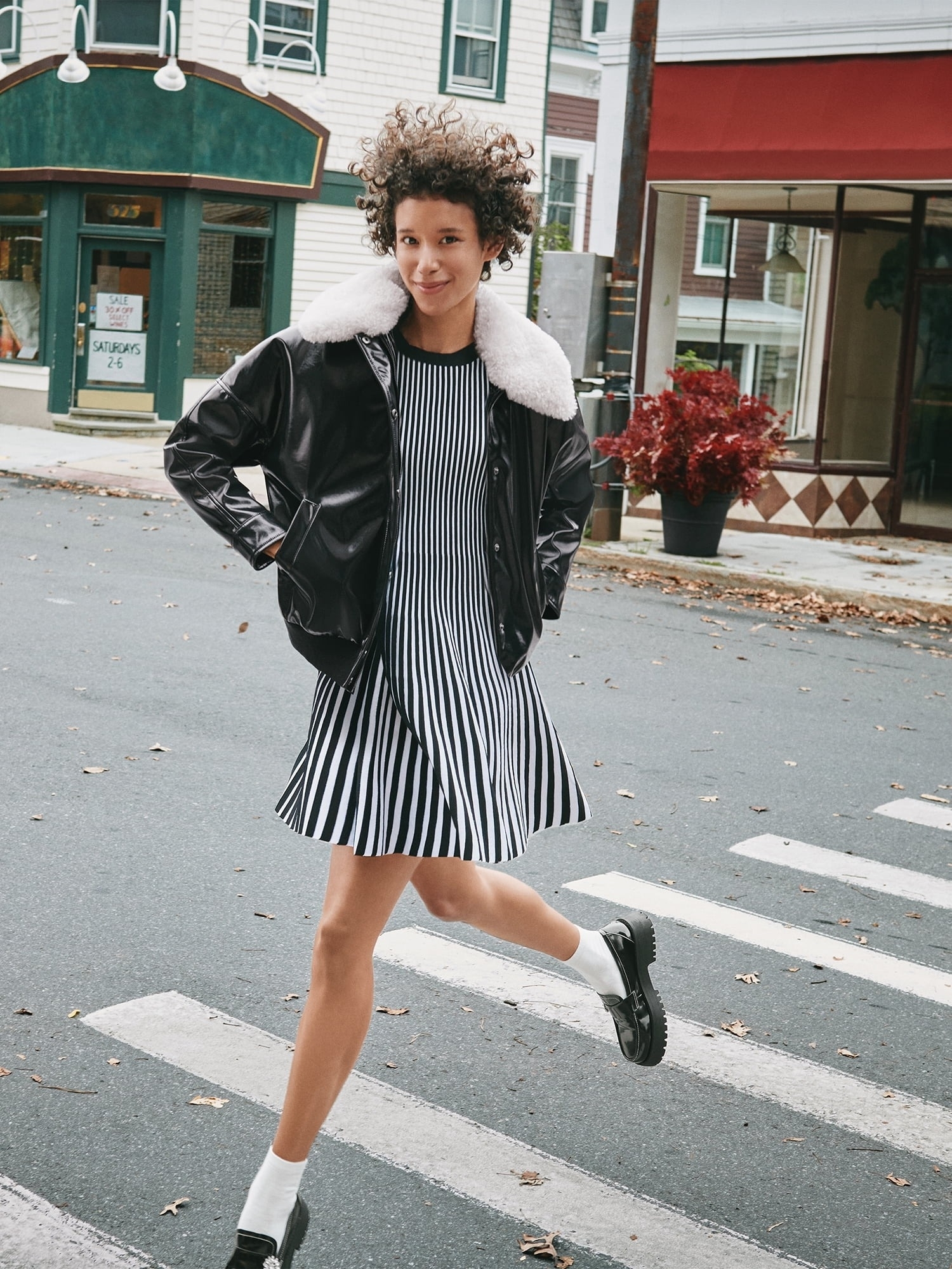 model in a striped dress and black jacket with fur collar, posing on a crosswalk