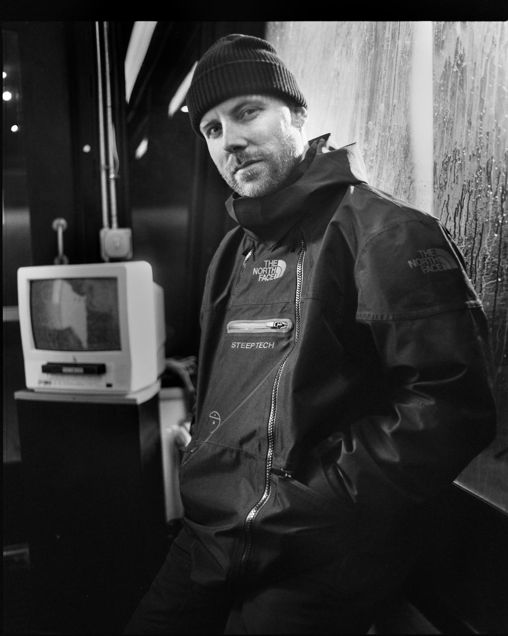 Man in a beanie and North Face jacket leaning against a wall with a vintage TV in the background
