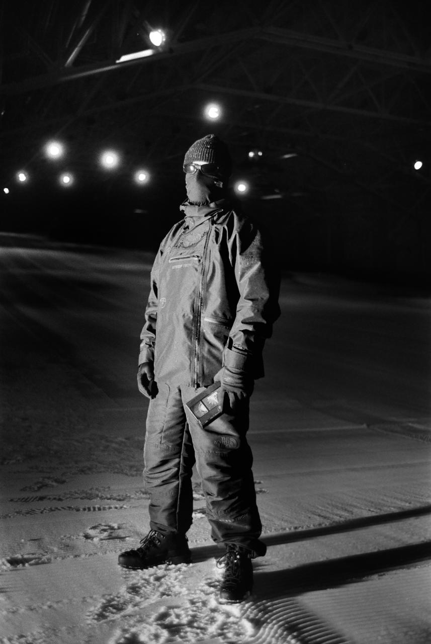 Person standing on snow at night under lights, wearing winter gear and a beanie, holding a book