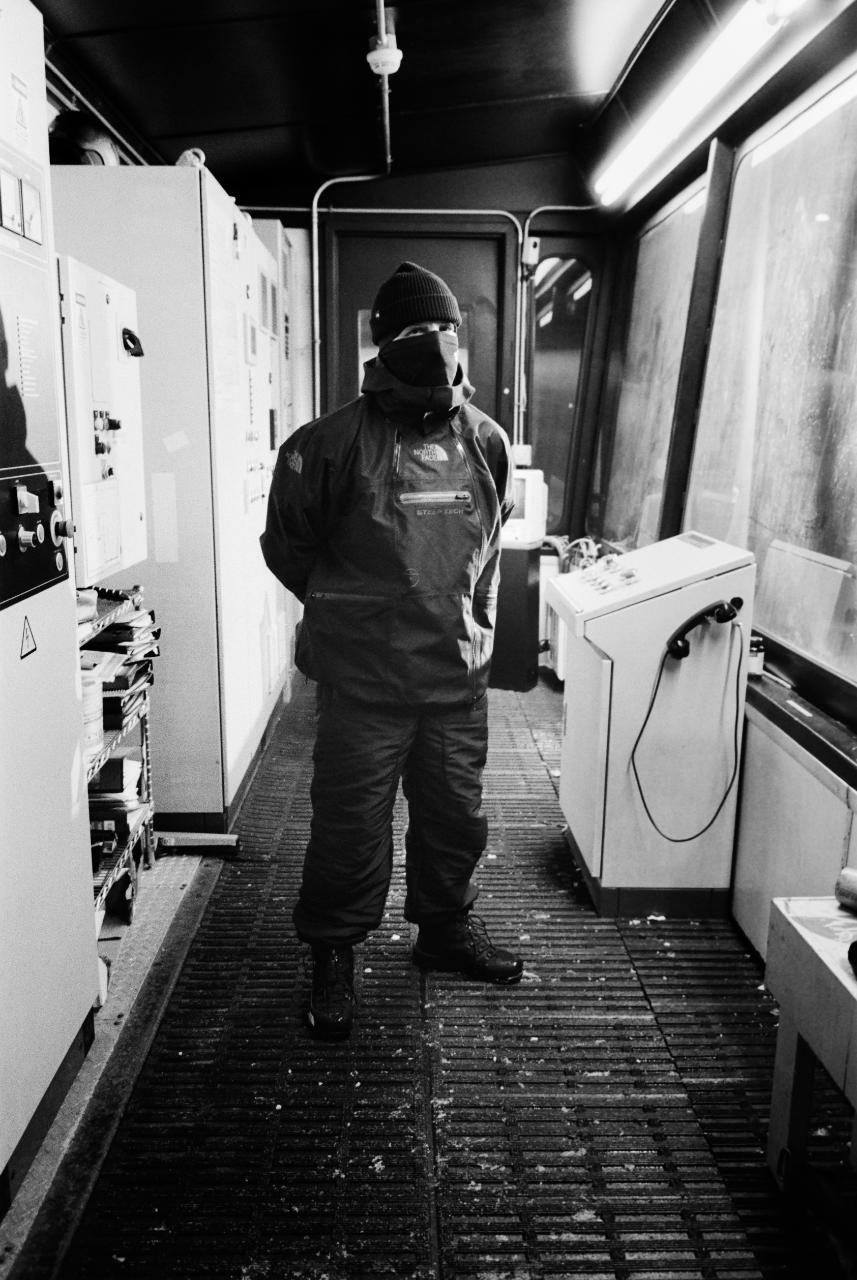Person in winter gear stands inside a room with electronic equipment and a washing machine
